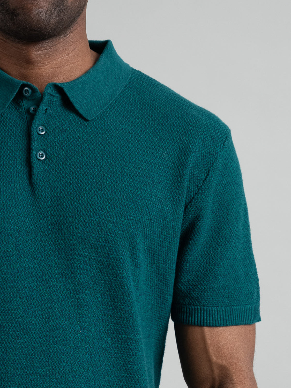 A person in a teal Austin Knitted Cotton Polo by ID Menswear, shown from shoulders to waist against a plain backdrop. Known for its breathable fit, the cotton-knit polo features a collar and three buttons. Only part of their face is visible.