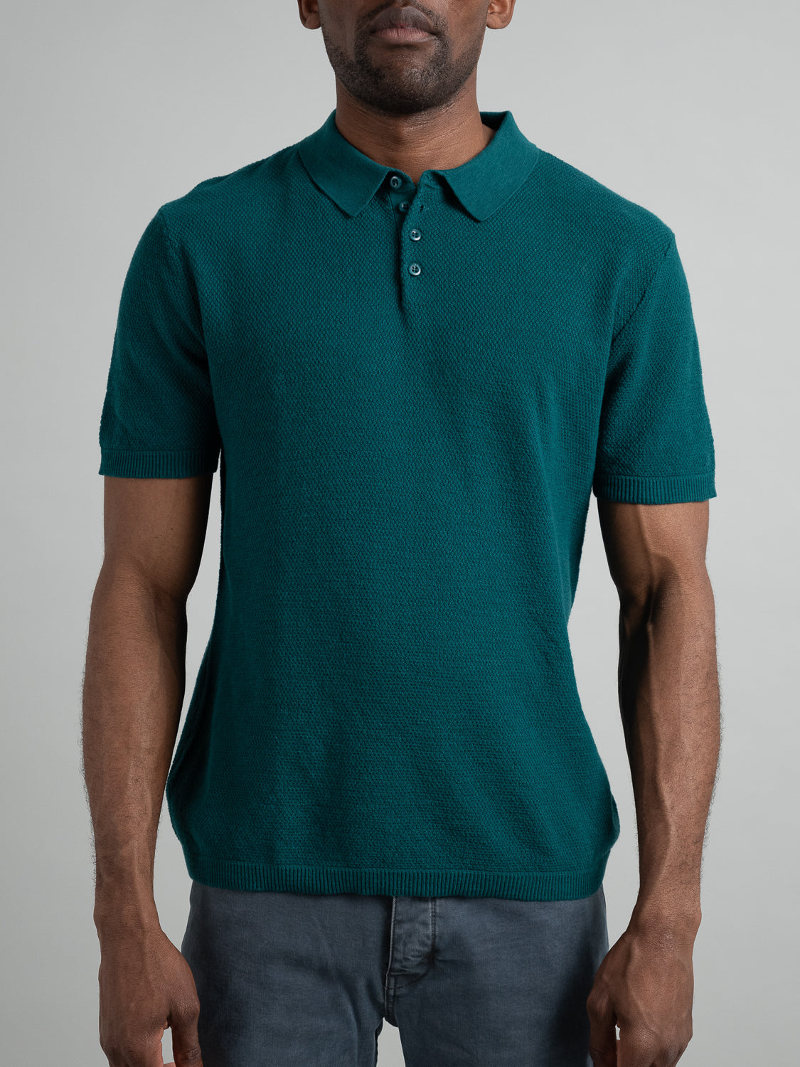 A man wears an Austin Knitted Cotton Polo from ID Menswear, a teal short-sleeve with a textured pattern, highlighting its breathable fit against a plain gray background.