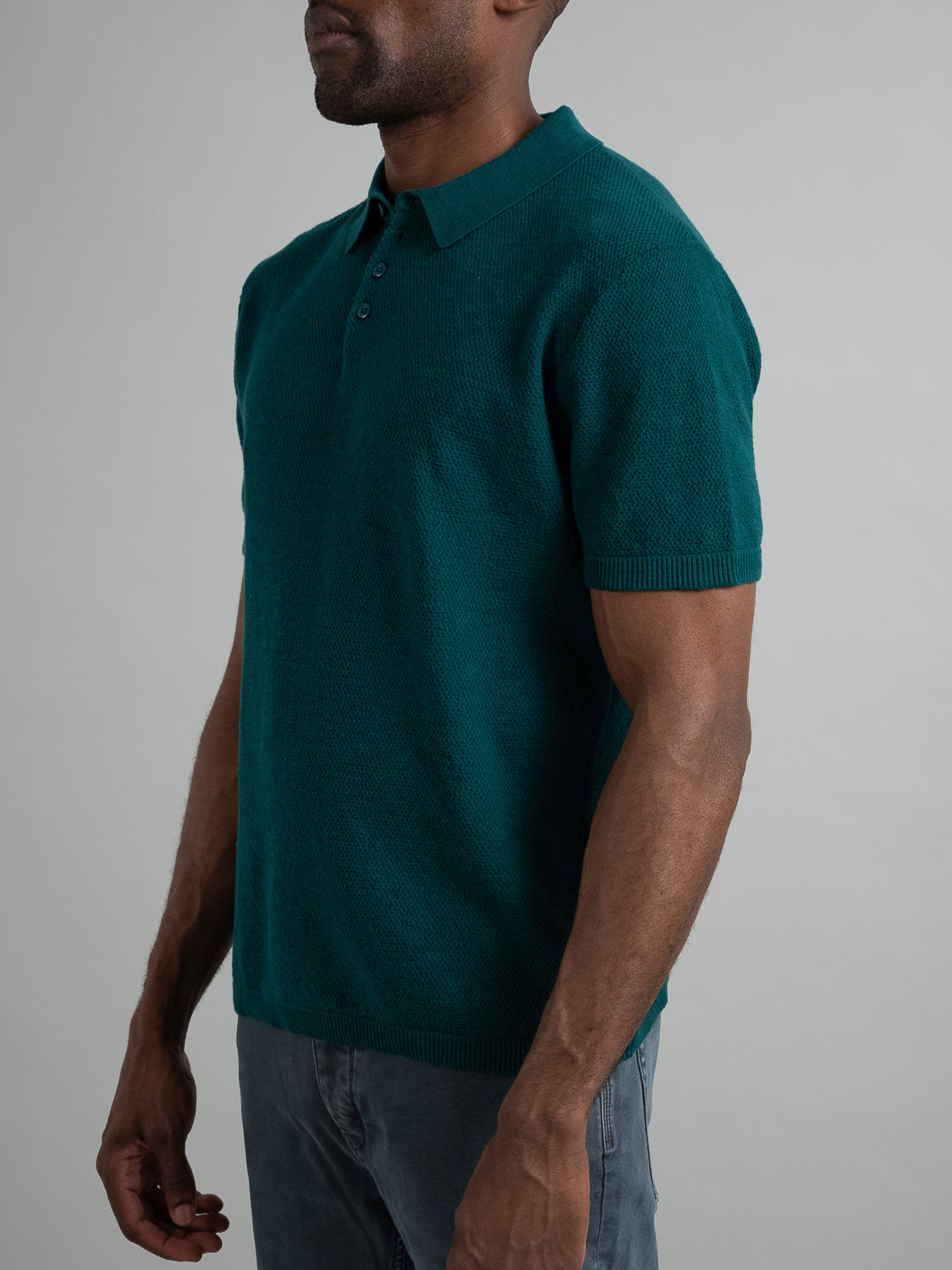 Someone stands against a plain background wearing the ID Menswear Austin Knitted Cotton Polo in teal and gray pants. The image captures the essence of casual wear, focusing on the upper body and part of the arms, with the head partially out of frame.