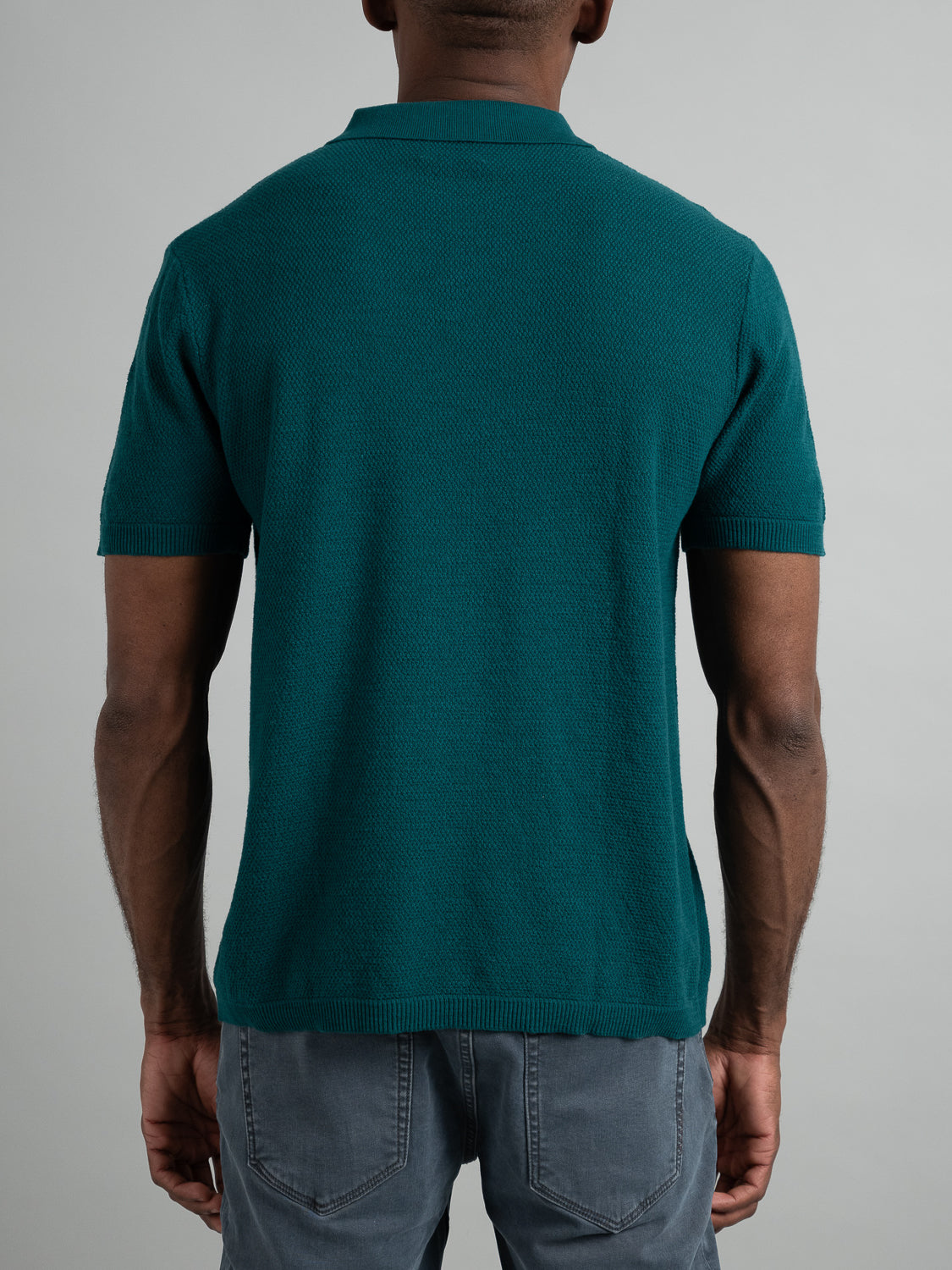 A person wears an ID Menswear Austin Knitted Cotton Polo in teal with gray pants, perfect for casual wear with a breathable fit, standing against a plain gray background, facing away from the camera.