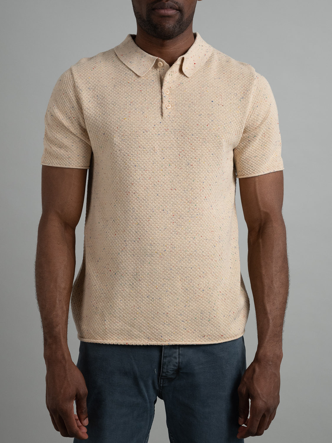 A man models a Denver Speckled Knit Cotton Polo by ID Menswear against a plain gray background. This beige textured short-sleeve shirt, with its subtle dot pattern and button-up collar, is made from breathable knit cotton and pairs effortlessly with dark jeans for casual wear.