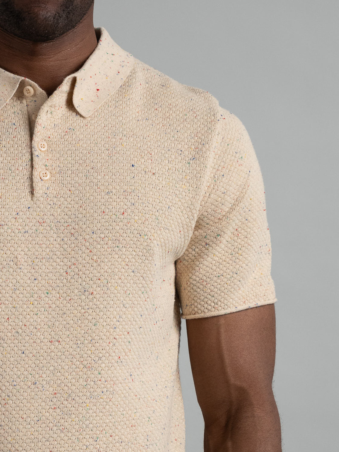 A person wears the Denver Speckled Knit Cotton Polo from ID Menswear, featuring a cream-color with multicolored speckles, short sleeves, buttons, and a collar. This cotton-knit polo offers a breathable fit ideal for casual outings against a plain gray backdrop.