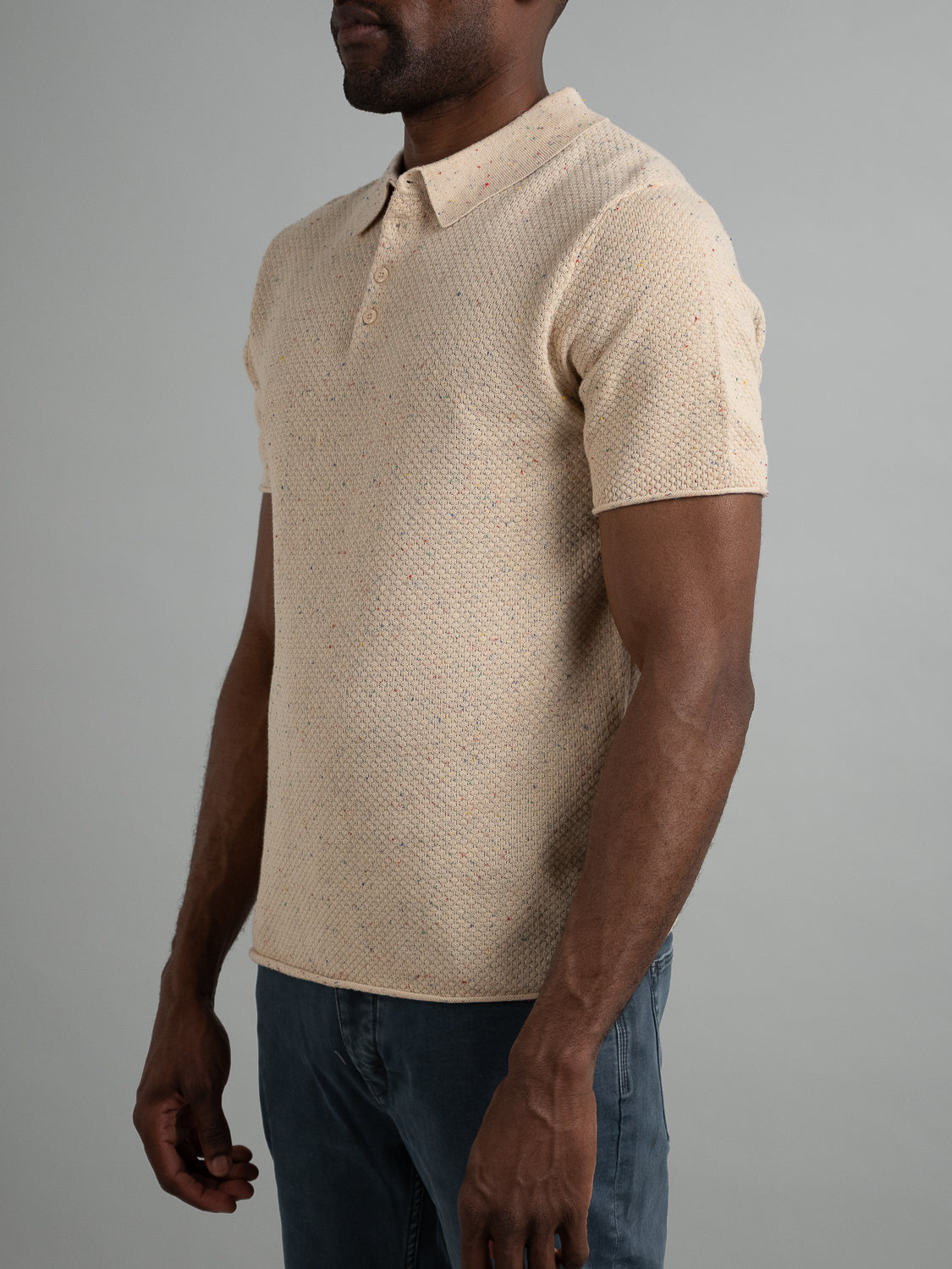 A person wearing the Denver Speckled Knit Cotton Polo by ID Menswear stands against a plain gray background. Paired with blue jeans, this beige polo exudes a relaxed style with breathable cotton-knit material, as arms rest comfortably at the sides.