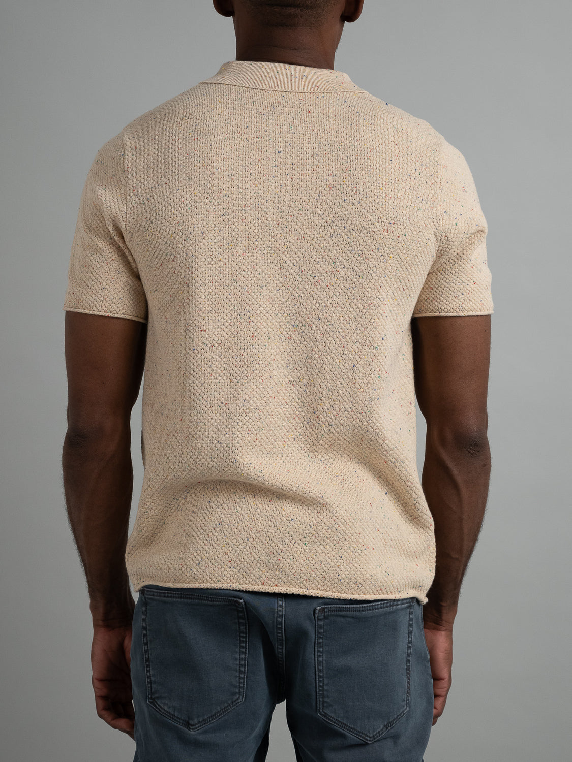 An individual is wearing the Denver Speckled Knit Cotton Polo from ID Menswear, featuring a light beige speckle pattern and short sleeves. The back view against a plain gray background highlights its breathable fit and casual style, complemented by blue jeans.