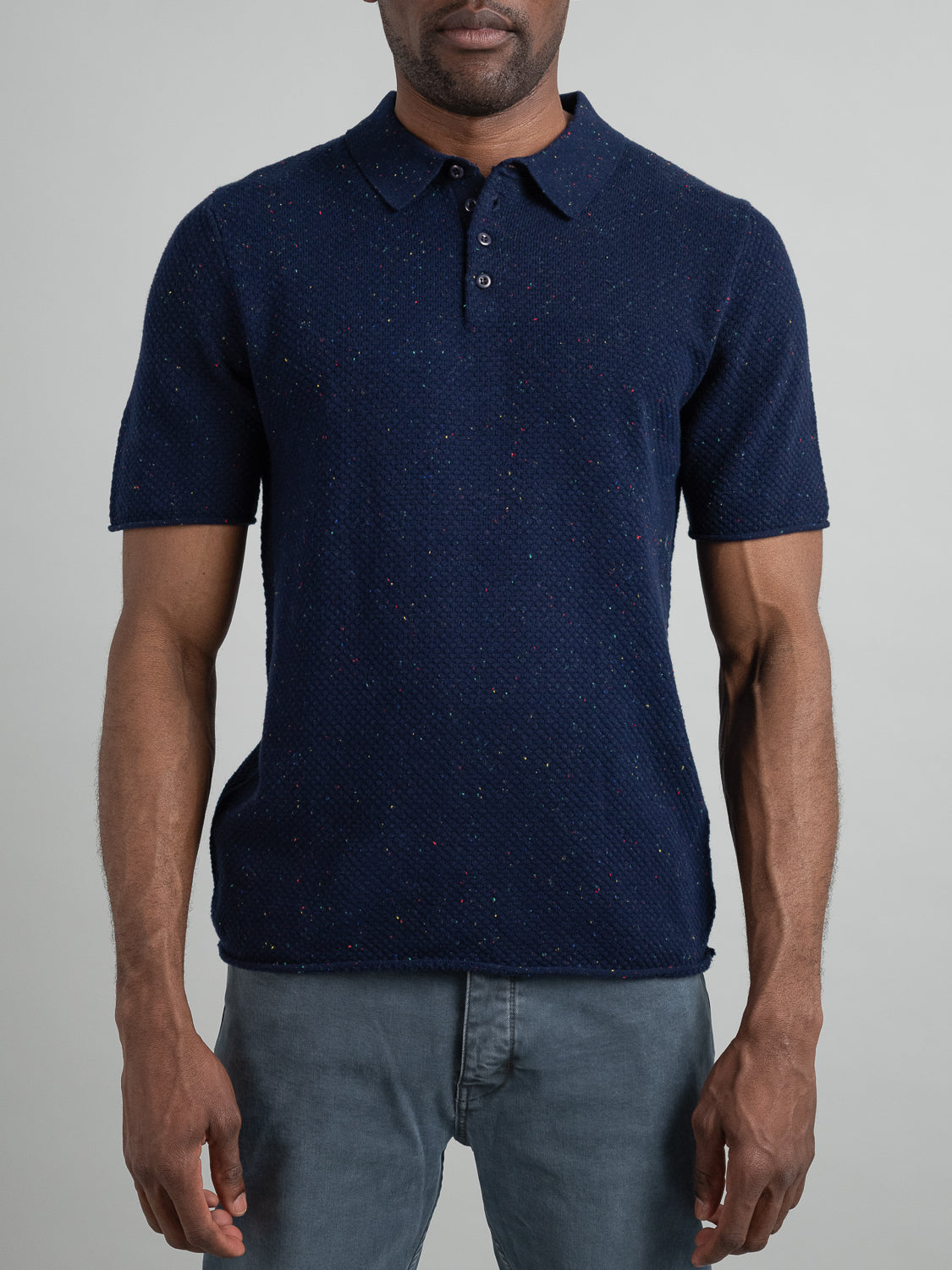 Standing against a plain gray background, a person wears the ID Menswear Denver Speckled Knit Cotton Polo, a dark blue, short-sleeved shirt with colorful speckles and textured knit pattern, paired with gray pants. Perfect for those seeking casual style and breathable comfort.