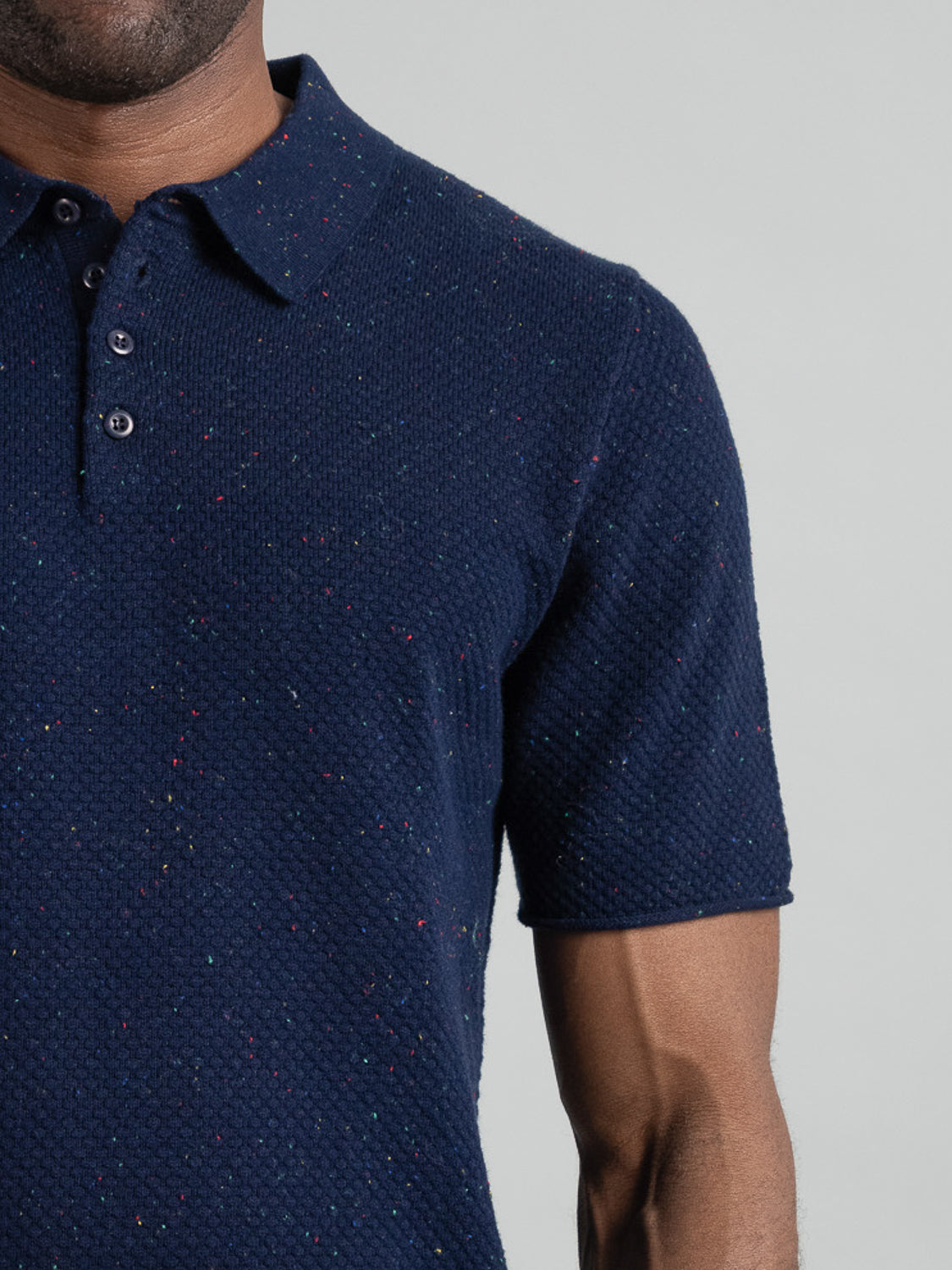 A person is wearing a Denver Speckled Knit Cotton Polo by ID Menswear. This dark blue, breathable cotton-knit polo has short sleeves, a speckled pattern, and three buttons at the collar. The background is plain light grey, showing only half of the persons torso.