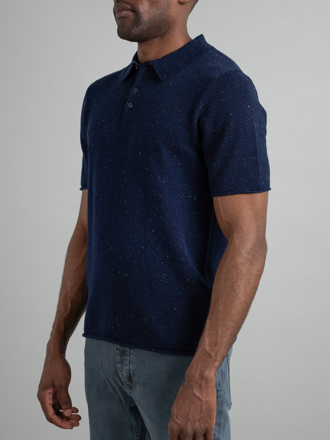 A person in the Denver Speckled Knit Cotton Polo by ID Menswear, featuring navy blue textured short sleeves and a buttoned collar, stands against a neutral background. Paired with light gray pants, this polo offers a breathable fit ideal for casual wear.