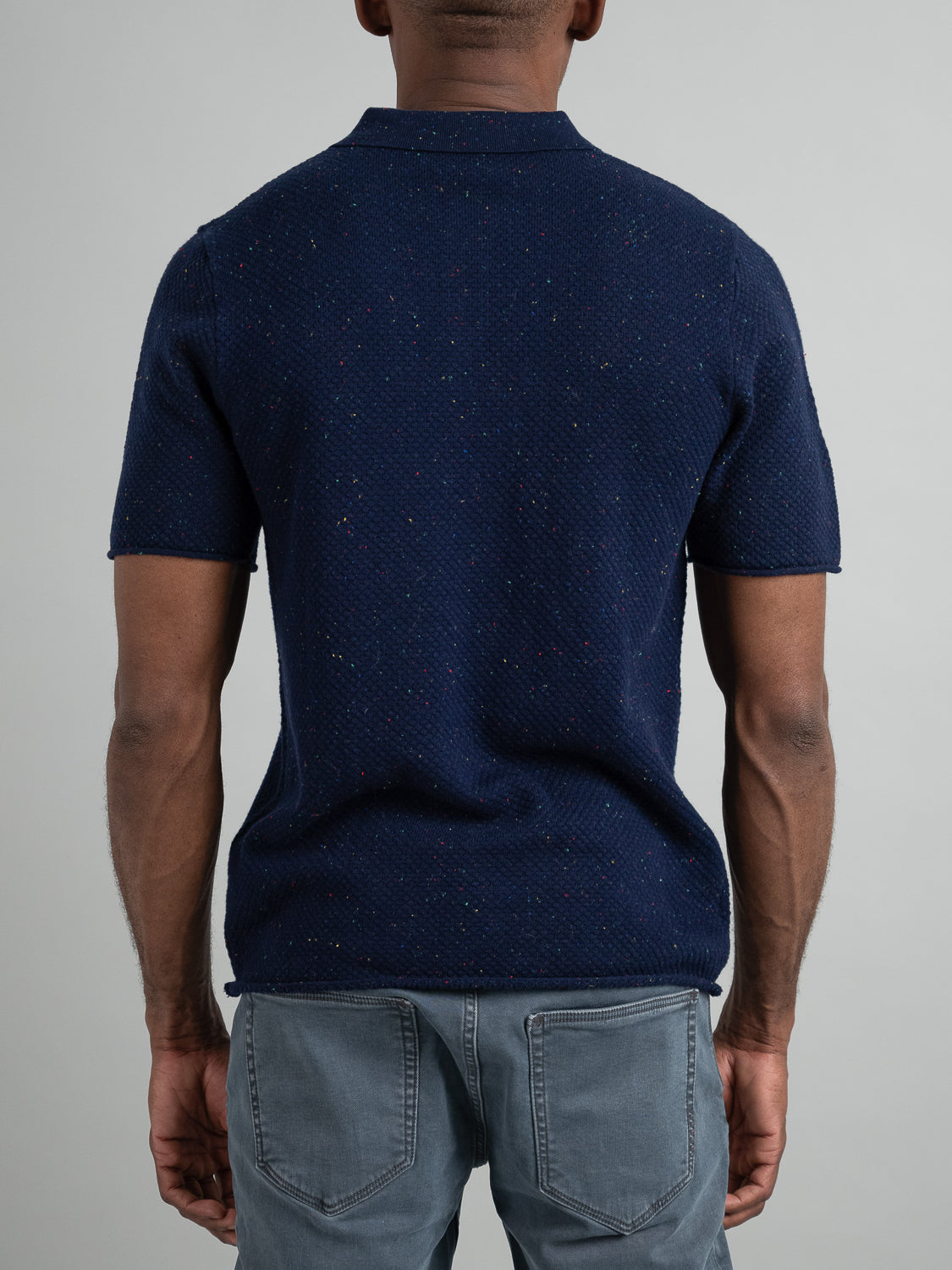 Facing away, a person wears the Denver Speckled Knit Cotton Polo by ID Menswear and light blue jeans. The breathable fit of the dark blue polo adds comfort to this casual look against a plain, light gray background.