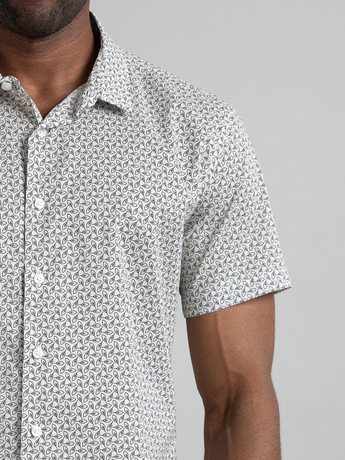 A person wears the neatly pressed Laurel Short Sleeve Cotton Printed Shirt by ID Menswear, featuring a small intricate black pattern on white. Against a plain gray backdrop, only the torso and part of the arm are visible.