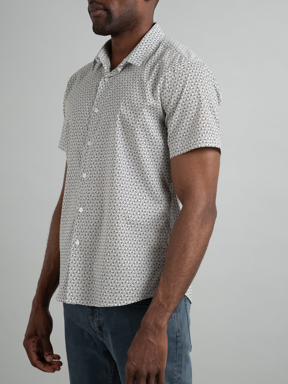 Someone wearing an ID Menswear Laurel Short Sleeve Cotton Printed Shirt stands against a plain background. The white shirt sports intricate black designs, paired stylishly with dark jeans. The image is cropped from the neck down.