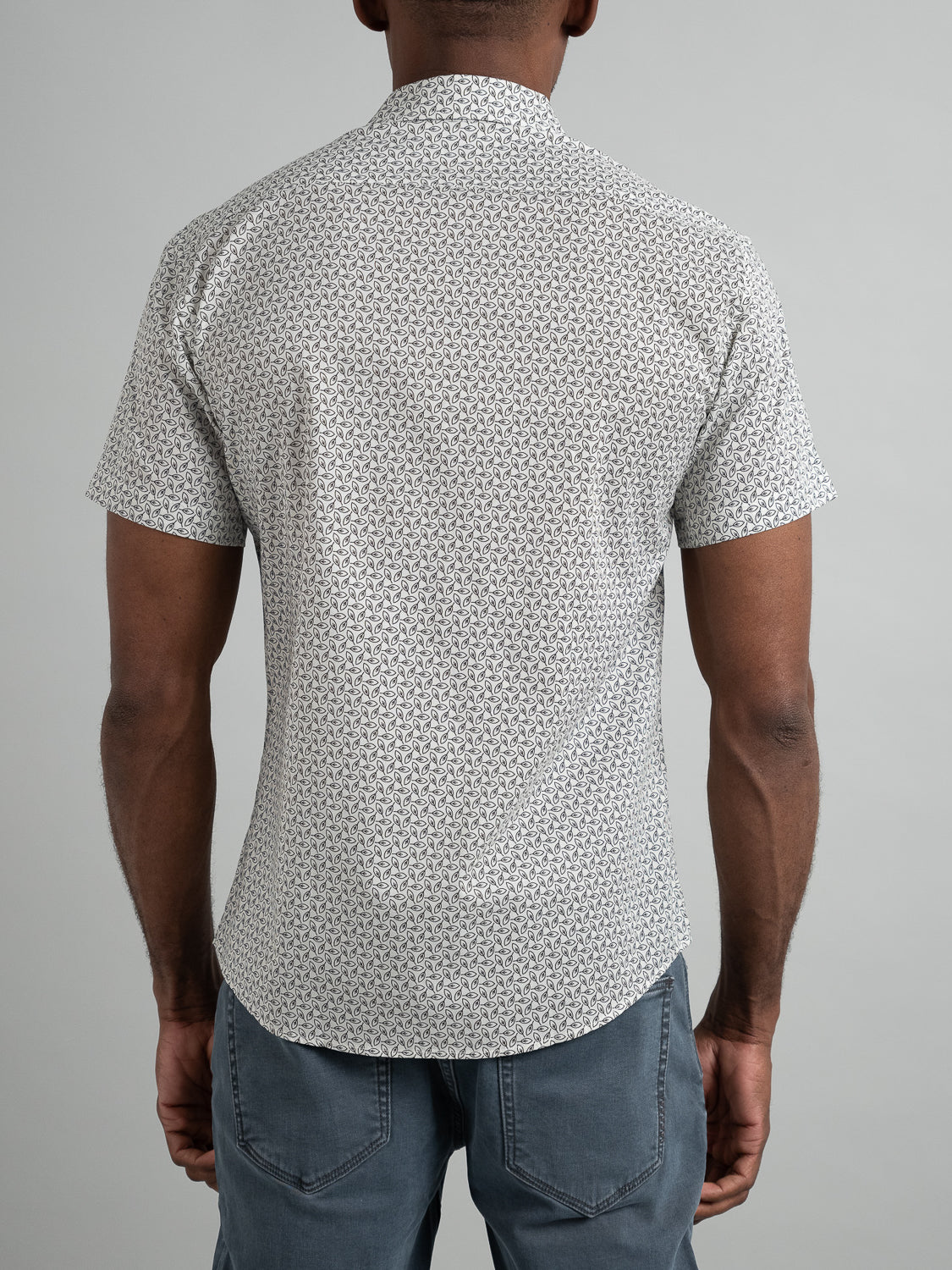 An individual with short hair, facing away, wears the Laurel Short Sleeve Cotton Printed Shirt by ID Menswear. The shirt has a subtle black and white geometric pattern and is paired with gray pants against a plain light gray backdrop.