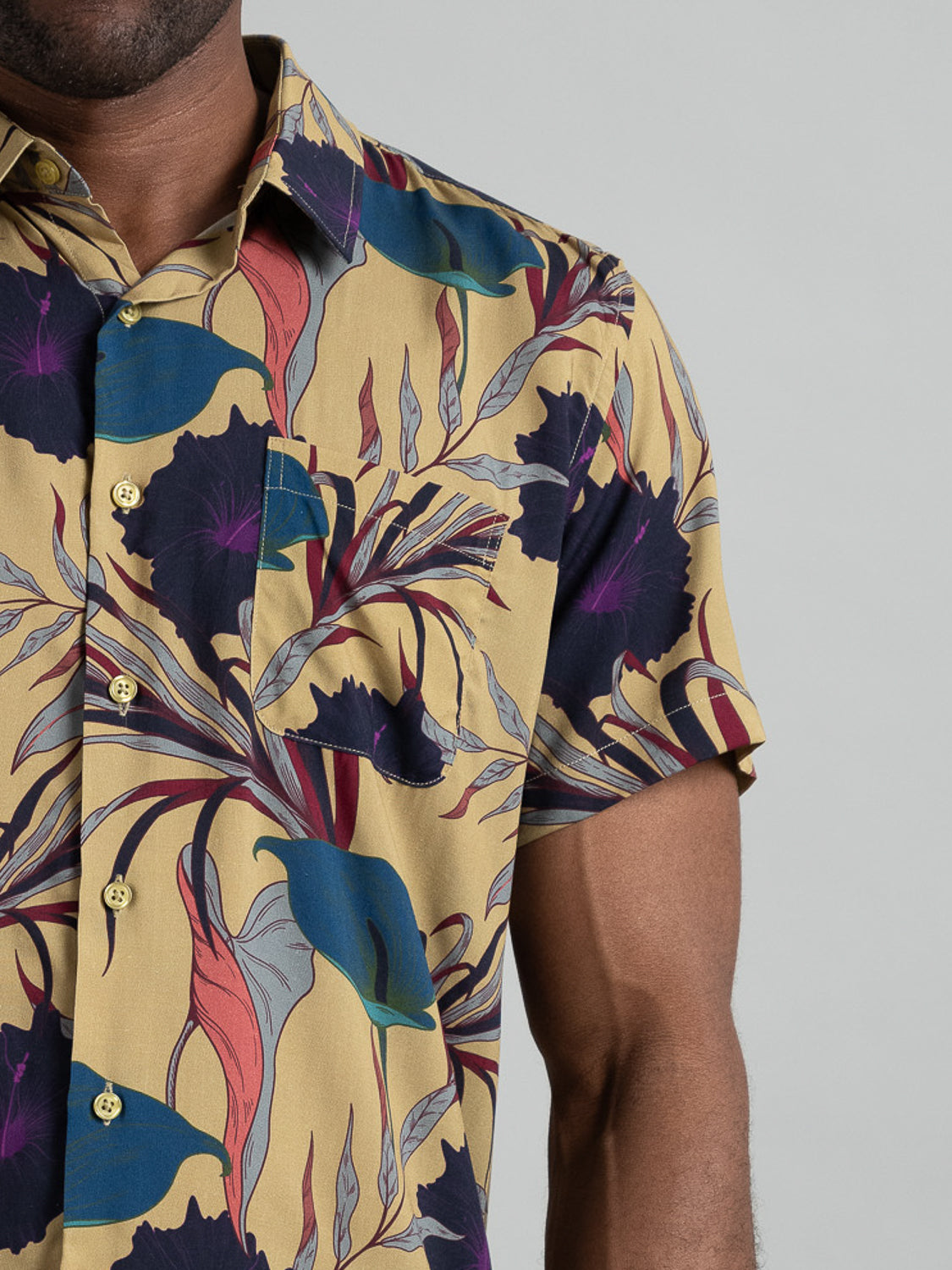 A person models ID Menswears Hibiscus Short Sleeve Printed Rayon Shirt, displaying vibrant blue, red, and purple floral prints on a sunny yellow background. Perfect for warm weather, the shirt includes a chest pocket while only part of the persons body is shown.