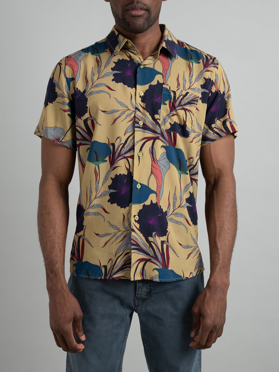 A person wears the ID Menswear Hibiscus Short Sleeve Printed Rayon Shirt, featuring a vibrant floral and leaf print on a beige background, paired with blue jeans against a light gray backdrop. This colorful button-up in shades of purple, blue, and red is ideal for warm weather.