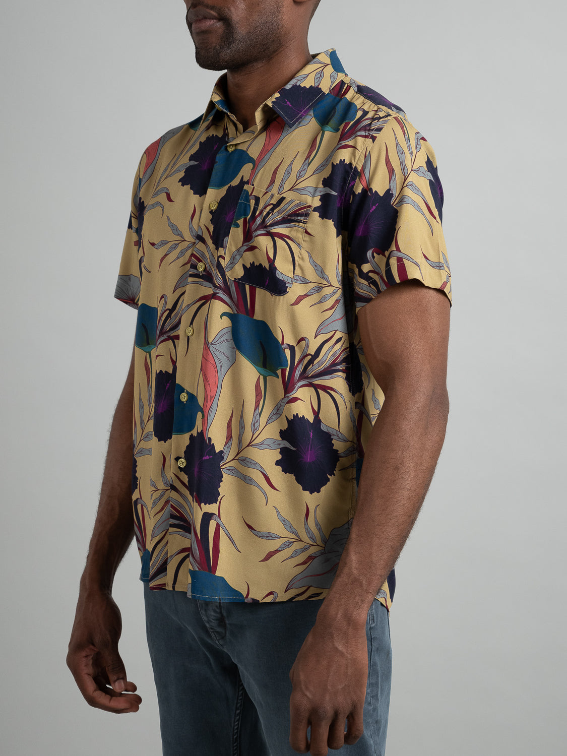 A man wears the Hibiscus Short Sleeve Printed Rayon Shirt from ID Menswear, featuring vibrant purple, teal, and mustard yellow floral prints on a beige backdrop. Ideal for warm weather, he pairs it with dark pants against a plain gray background.