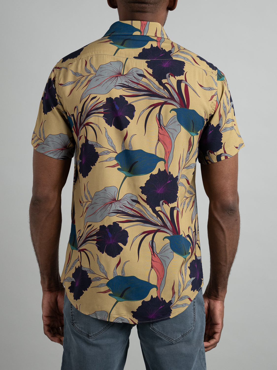 A person wearing the ID Menswear Hibiscus Short Sleeve Printed Rayon Shirt in vibrant blue, purple, and red on tan is seen from the back. They pair it with gray jeans for a stylish warm-weather look.