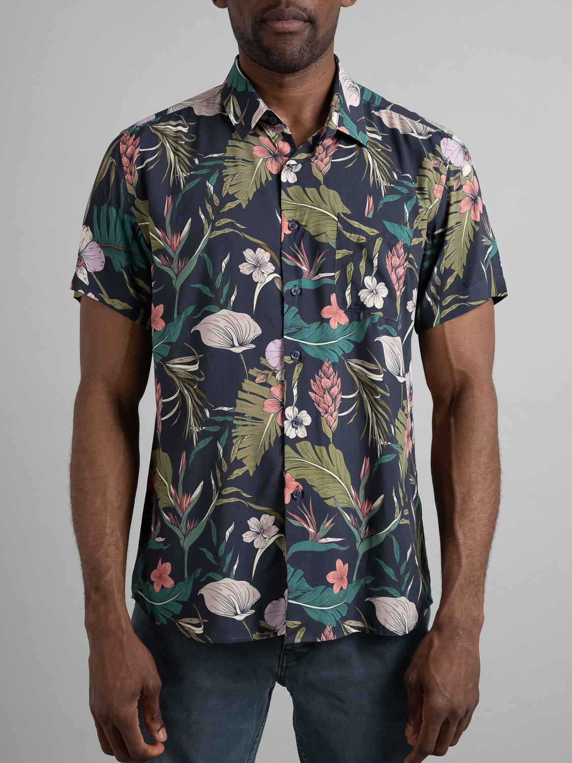 Someone is wearing the Kauai Short Sleeve Printed Rayon Shirt from ID Menswear, featuring colorful floral prints of green leaves, and pink, white, and red flowers. The vibrant shirt is paired with dark pants against a plain light background.