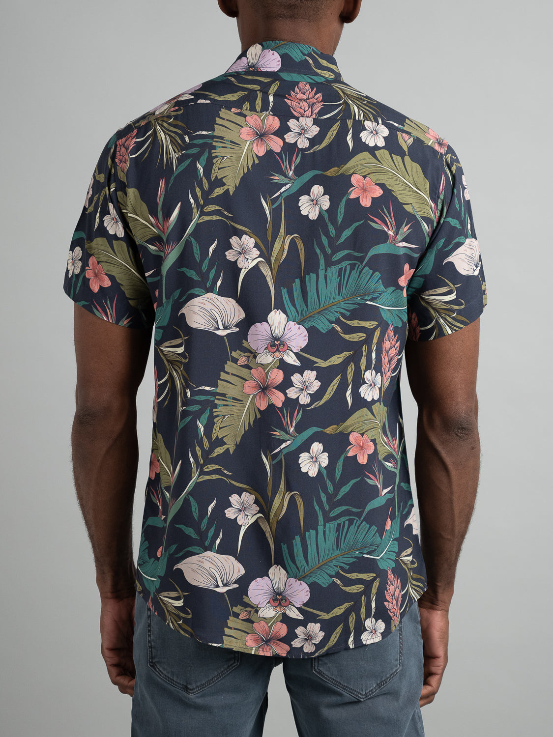 Against a plain background, someone wearing ID Menswears Kauai Short Sleeve Printed Rayon Shirt, featuring vibrant floral and leaf patterns in dark tones, stands with their back to the camera, paired with light blue jeans.