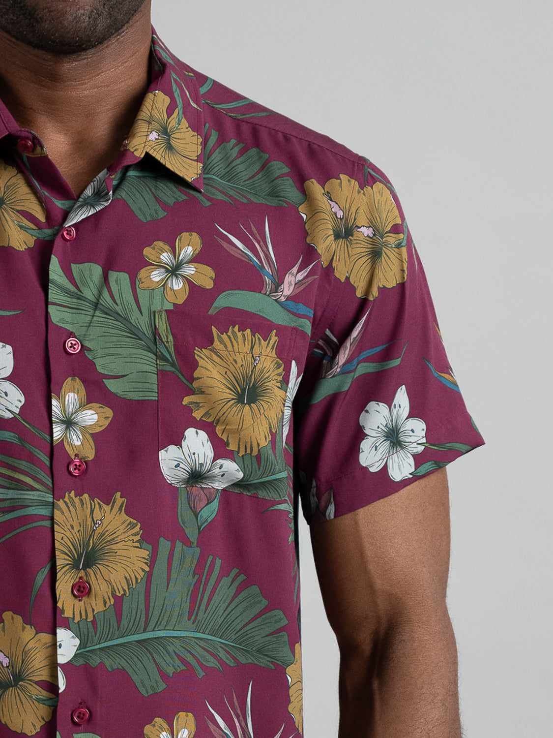 A person wears the Bali Short Sleeve Printed Rayon Shirt from ID Menswear, showcasing a maroon base with vibrant white and yellow floral prints, green leaves, dark red buttons, and a collar. Ideal for warm days, it features a plain light gray background.