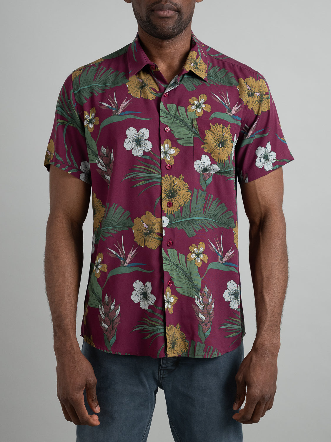 A person in ID Menswears Bali Short Sleeve Printed Rayon Shirt, featuring vibrant maroon fabric adorned with yellow and white flowers and green leaves, paired with blue jeans on a plain background. Ideal for warm weather, the persons face is not visible.