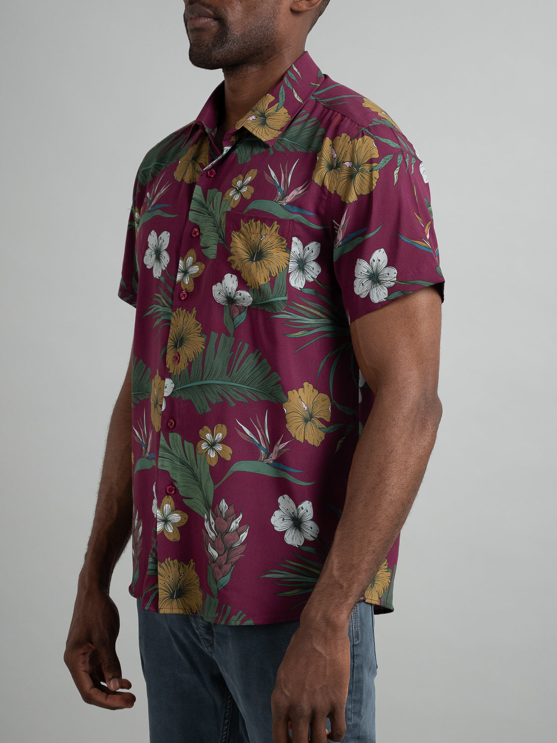 A man wears the ID Menswear Bali Short Sleeve Printed Rayon Shirt featuring vibrant floral prints in yellow, white, and pink with green leaves. The maroon Hawaiian shirt is perfect for warm weather, and his dark skin contrasts elegantly against the plain gray background.