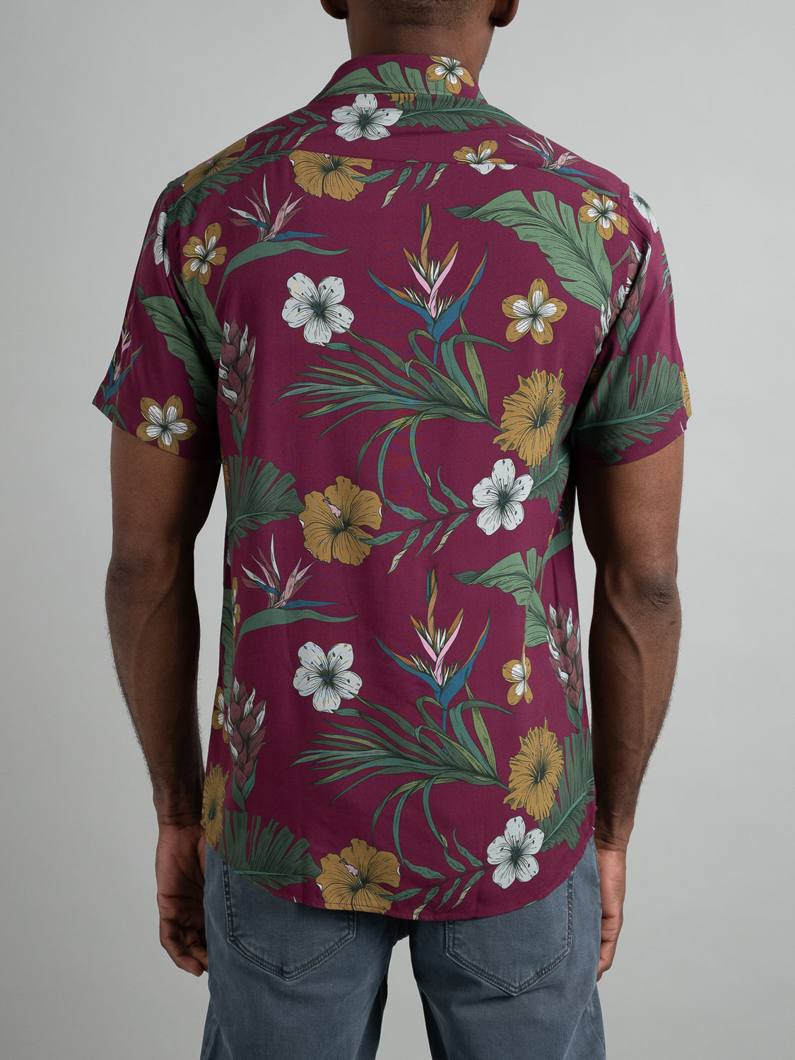 An individual wears the Bali Short Sleeve Printed Rayon Shirt by ID Menswear, featuring maroon fabric with vibrant yellow, white, and green floral prints. Its paired with gray pants on a plain background, making it ideal for warm weather days.