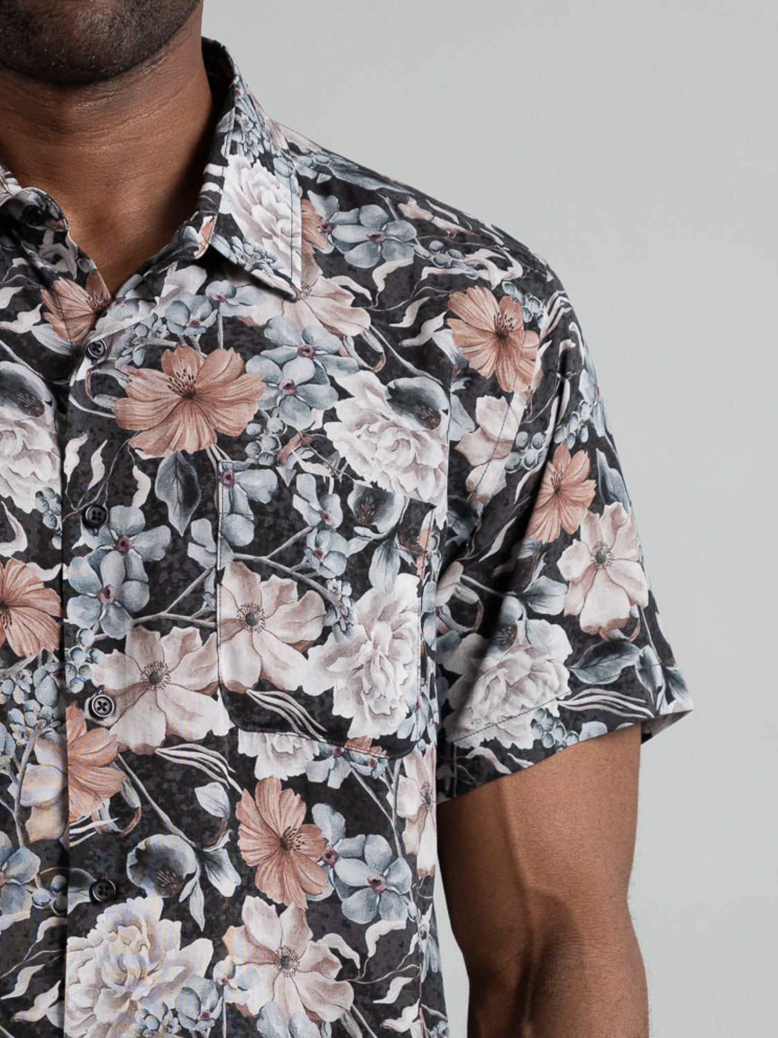 Close-up of a person wearing the Tobe Short Sleeve Printed Rayon Shirt by ID Menswear, showcasing vibrant floral prints in black, gray, and dusty pink against a plain gray background—ideal for embracing warm weather vibes.