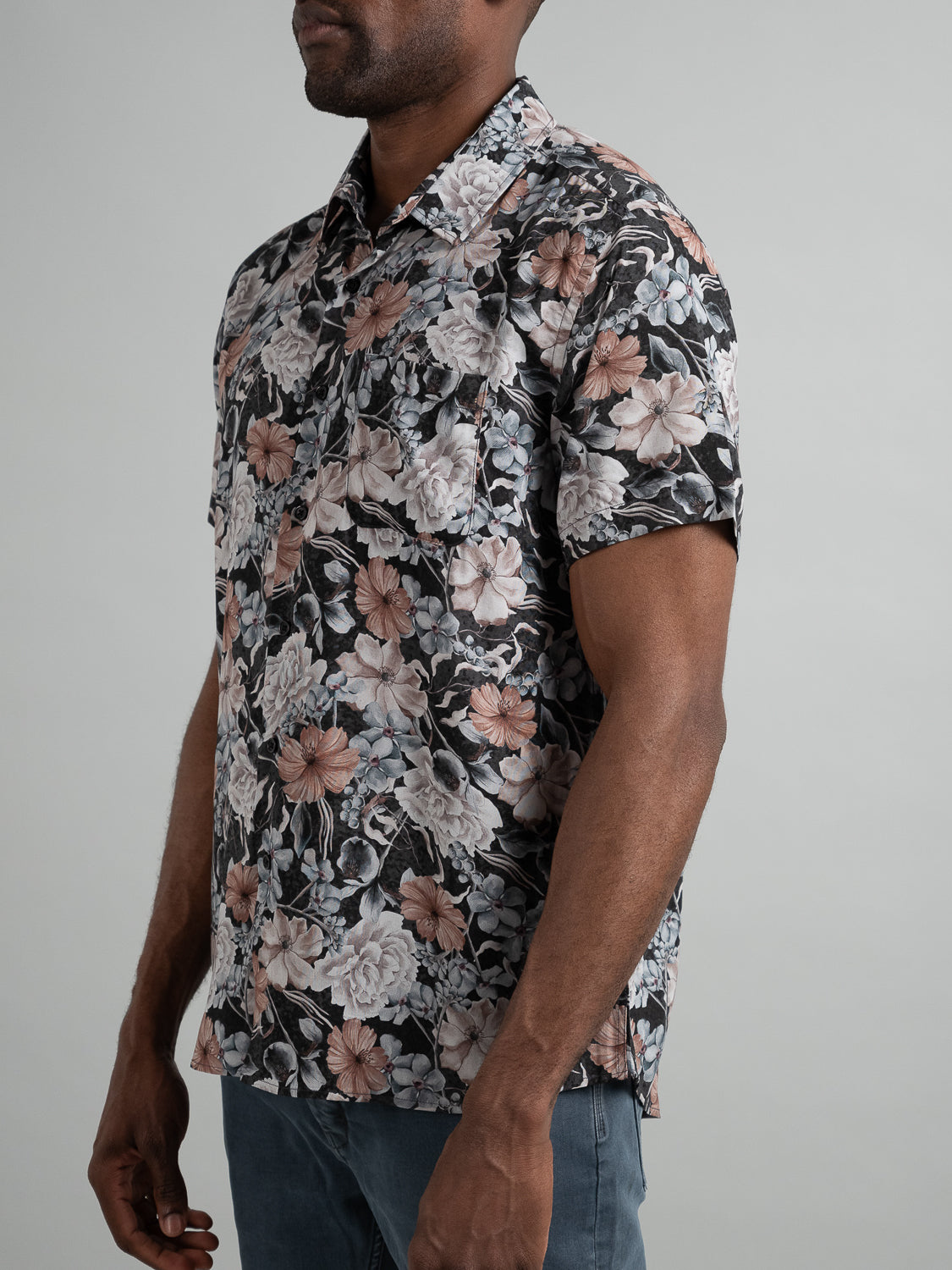 A person in an ID Menswear Tobe Short Sleeve Printed Rayon Shirt, featuring vibrant pink and gray floral patterns, stands against a plain background. Paired with blue jeans, they are seen from the side, looking away from the camera. Ideal for warm weather.