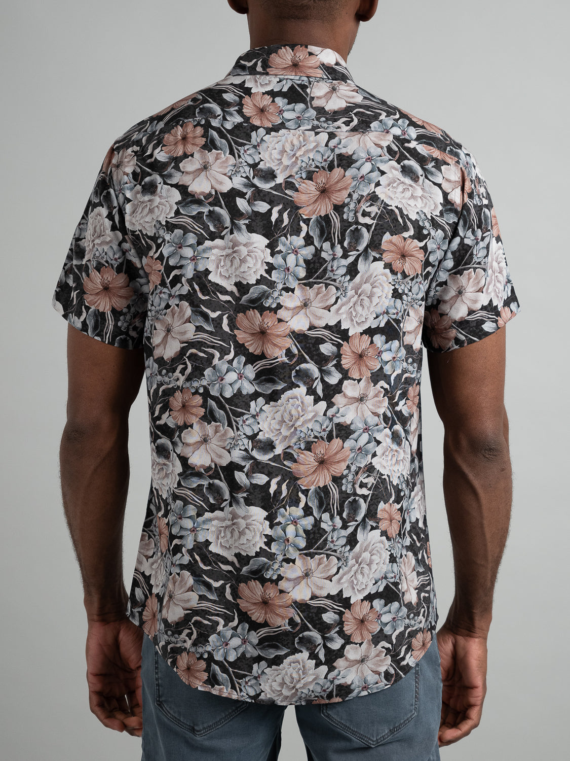 Someone stands with their back to the camera, wearing ID Menswears Tobe Short Sleeve Printed Rayon Shirt. This warm-weather style features large floral prints in pink, white, and gray on a dark background. Its effortlessly paired with blue jeans.