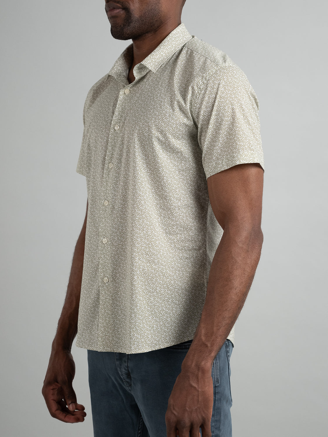A man models the ID Menswear Anise Short Sleeve Cotton Printed Shirt, featuring light patterns, against a plain gray backdrop. His face remains obscured for anonymity, and he completes the look with dark pants.