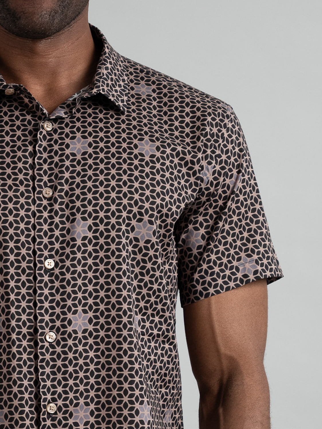 A person models the Petals Short Sleeve Cotton Printed Shirt from ID Menswear, featuring a black and pink geometric print with a collar and front buttons, set against a plain gray background. Only the upper half of their body is visible.