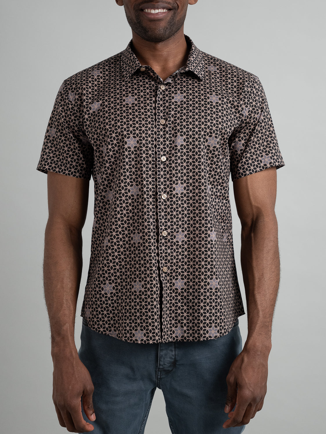 A person wearing ID Menswears Petals Short Sleeve Cotton Printed Shirt featuring a black and white geometric pattern is paired with dark blue jeans, set against a plain, light gray background.