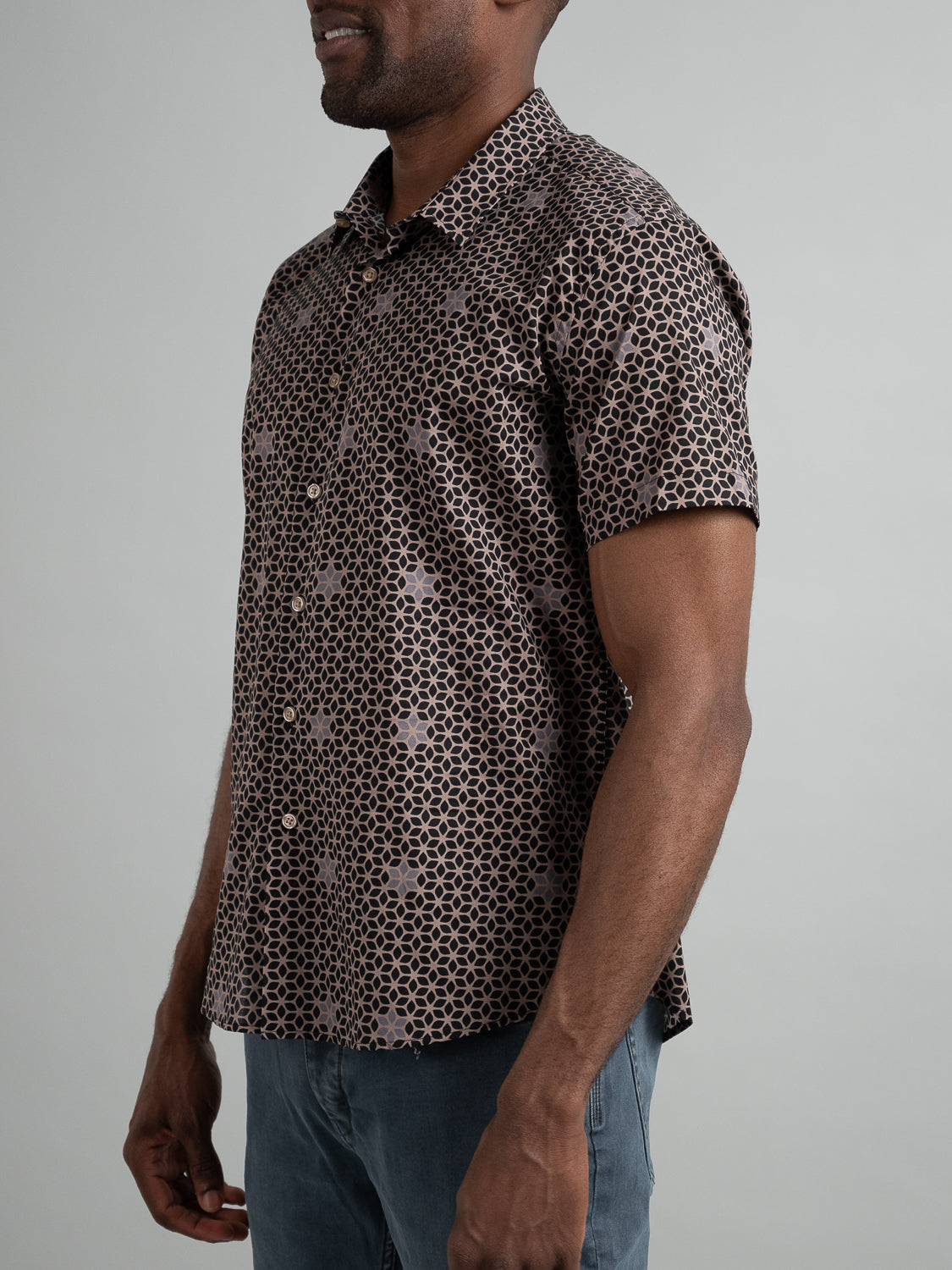 A person wears the ID Menswear Petals Short Sleeve Cotton Printed Shirt featuring a black and light brown geometric pattern, paired with light blue pants. The side view photo against a plain gray background highlights the shirts elegant lightweight design.