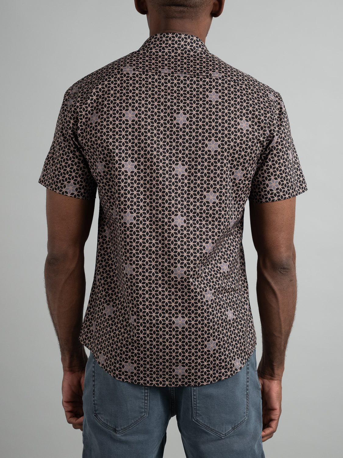 A person is seen from behind wearing an ID Menswear Petals Short Sleeve Cotton Printed Shirt with geometric black and brown patterns, paired with light gray pants against a plain, light-colored background.