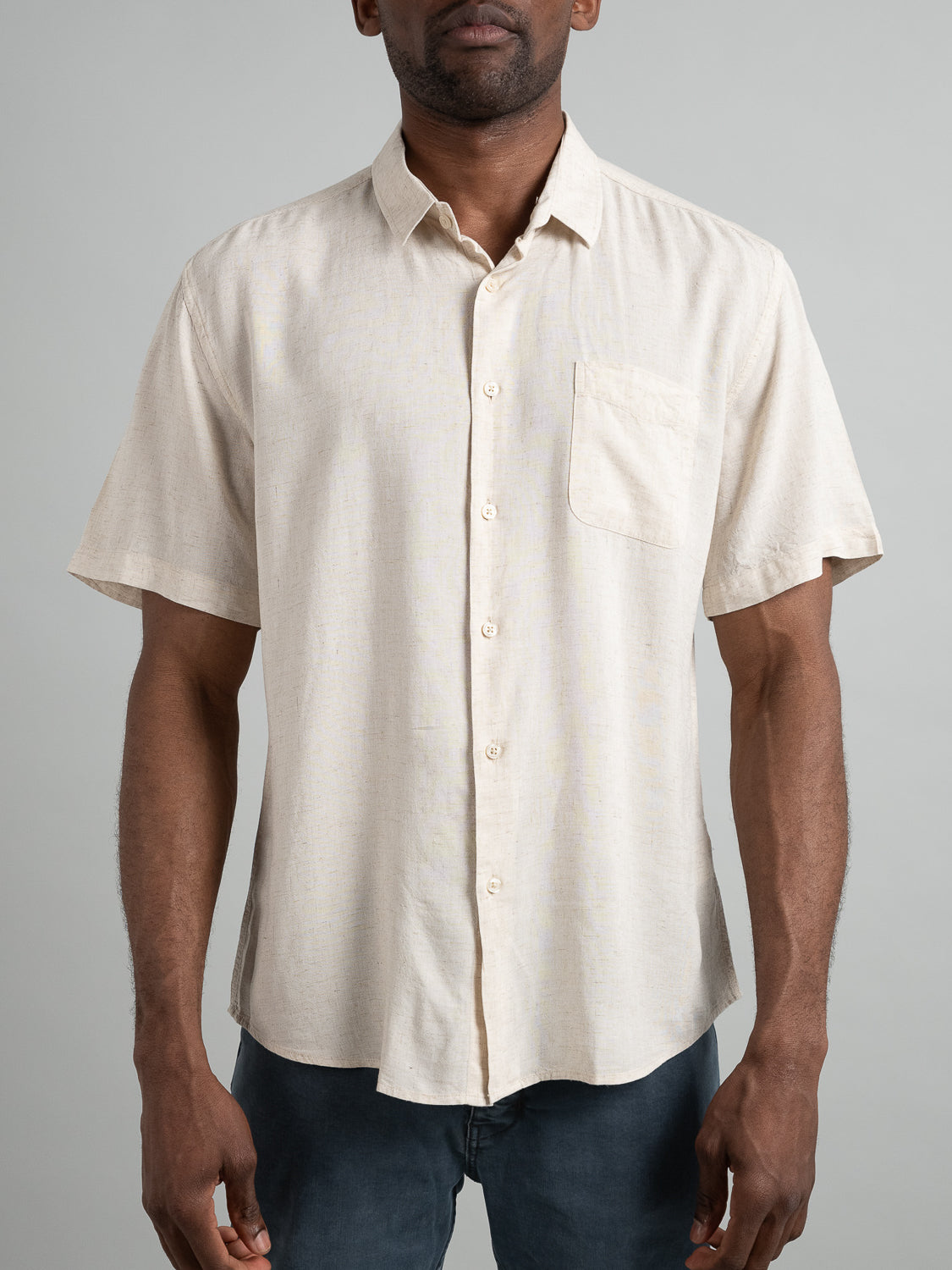 A person in a Maharishi Rayon Linen Short Sleeve Shirt from ID Menswear, featuring a light beige color, button-up style, and front pocket, stands against a plain gray background with hands at their sides. The shirt exudes both comfort and style.
