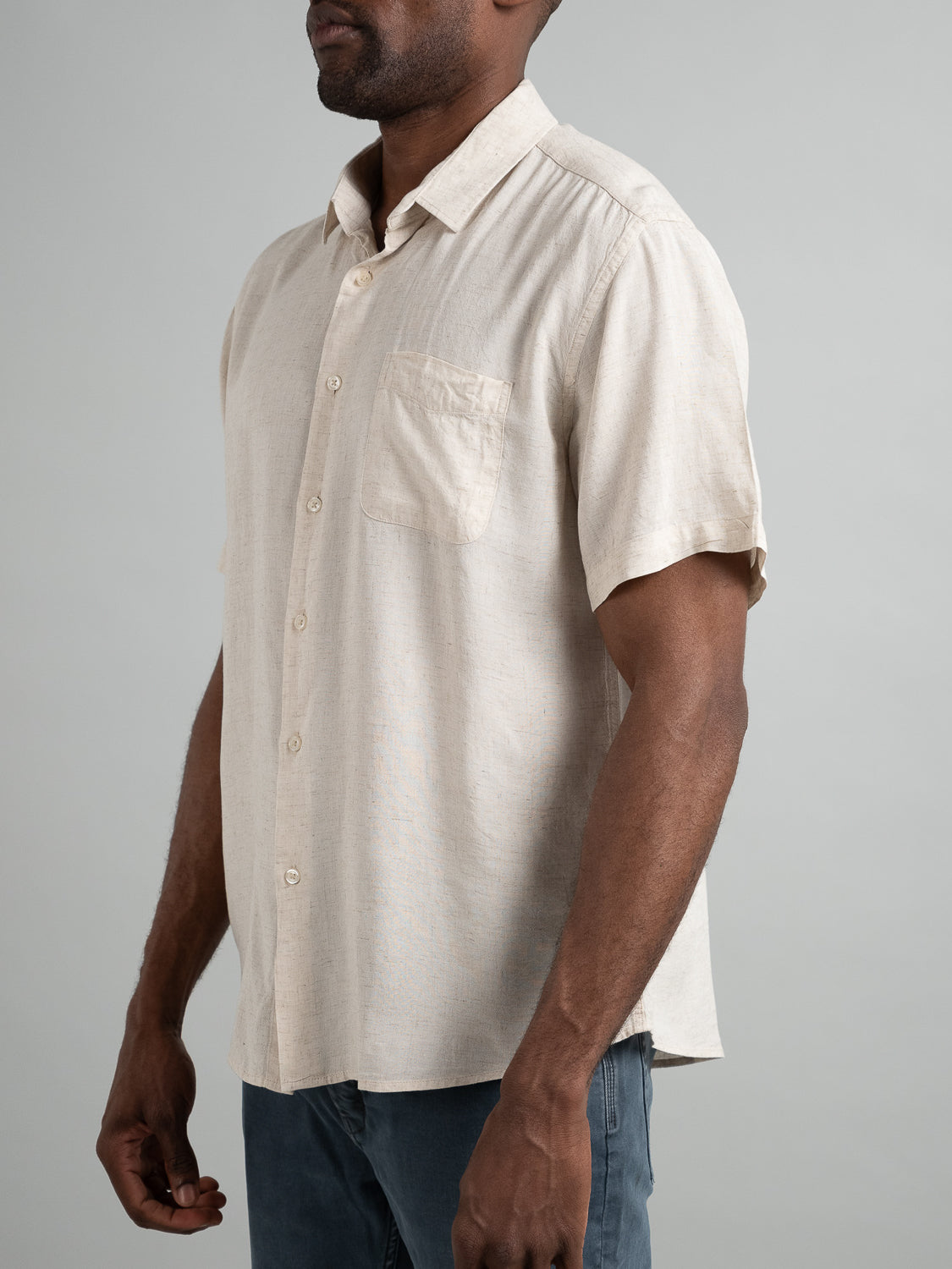 Against a gray backdrop, a person models the beige short-sleeve Maharishi Rayon Linen Shirt by ID Menswear, which features a front pocket. Their relaxed hands complement the casual look paired with blue jeans.