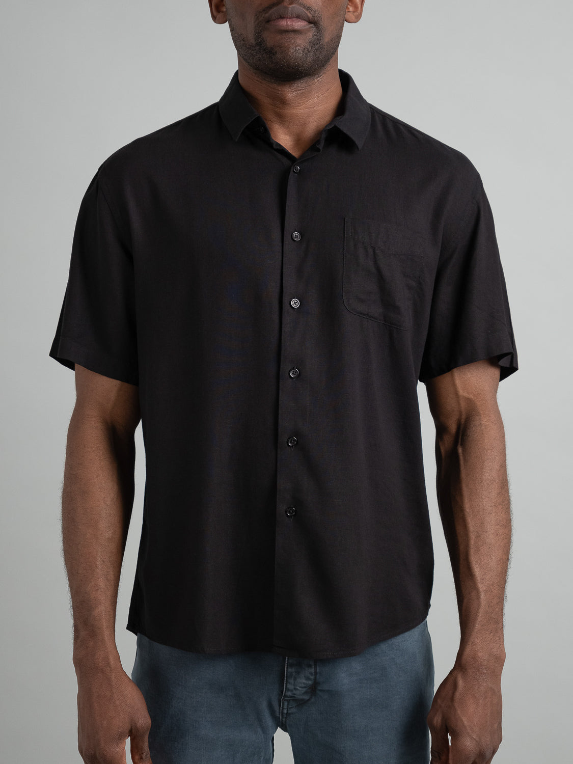 A person is wearing an ID Menswear Maharishi Rayon Linen Short Sleeve Shirt in black with a front pocket, paired with dark gray pants against a plain light gray background that enhances the stylish look.