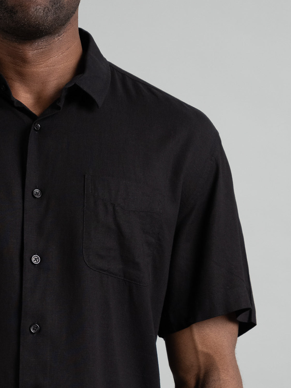Close-up of an individual donning the Maharishi Rayon Linen Short Sleeve Shirt by ID Menswear. The black shirt, featuring short sleeves and a chest pocket, is made from a rayon-linen blend for comfort and style against a plain, light grey background.