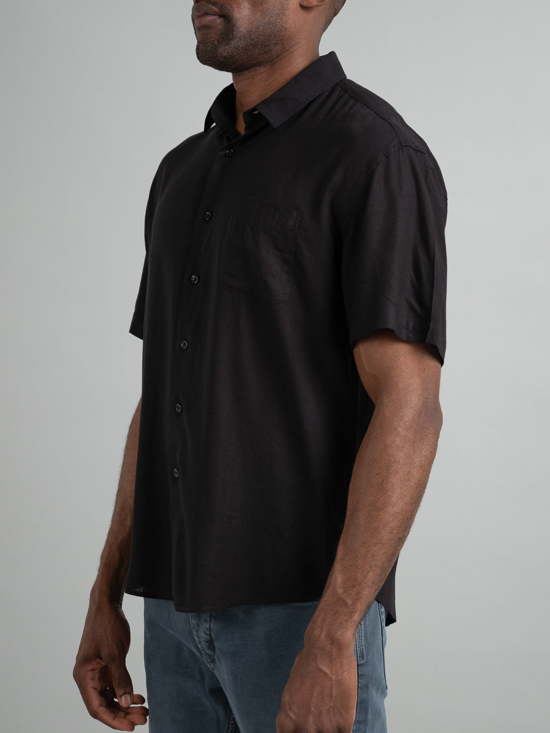 A person in a black ID Menswear Maharishi Rayon Linen Short Sleeve Shirt and blue jeans stands against a neutral background. The lightweight shirt features a chest pocket, with their hands relaxed at their sides.