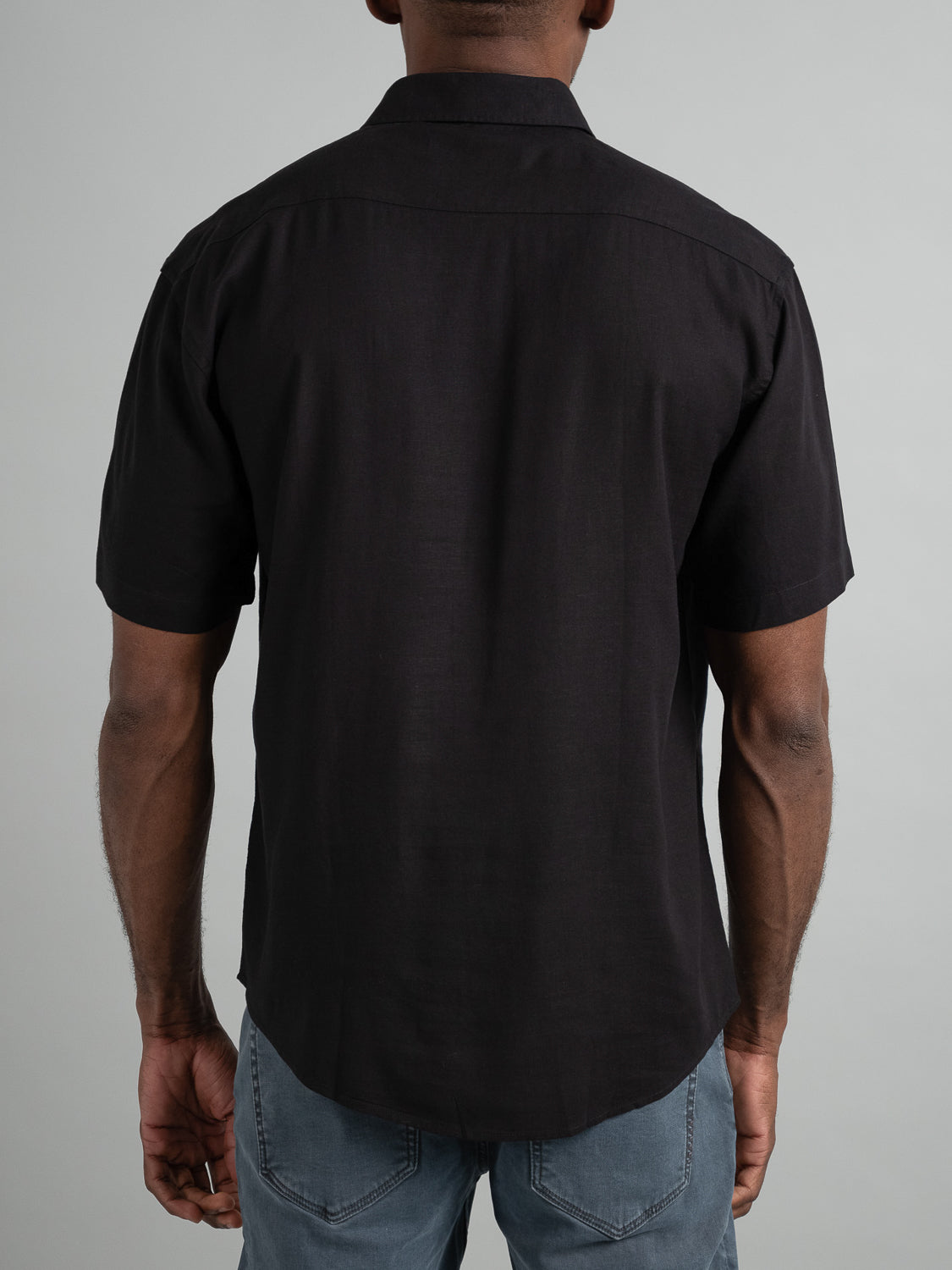 Rear view of a person wearing an ID Menswear Maharishi Rayon Linen Short Sleeve Shirt, its sleek black fabric draping effortlessly over blue jeans, standing against a plain gray background.