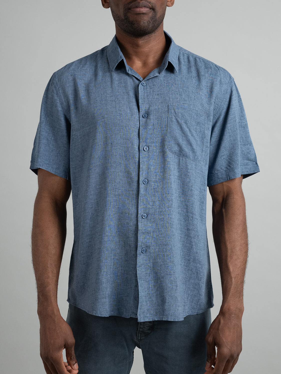A person wears the Maharishi Rayon Linen Short Sleeve Shirt by ID Menswear, a lightweight, short-sleeve button-up with a subtle textured pattern and left chest pocket, standing against a plain background.