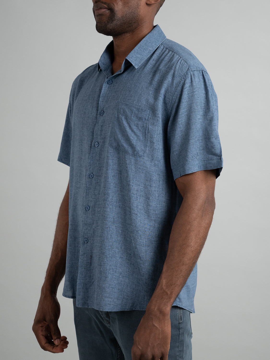 Someone is wearing a short-sleeved, button-up blue Maharishi Rayon Linen Shirt by ID Menswear featuring a left pocket, teamed with gray pants. The lightweight shirt ensures comfort against the plain light gray backdrop.