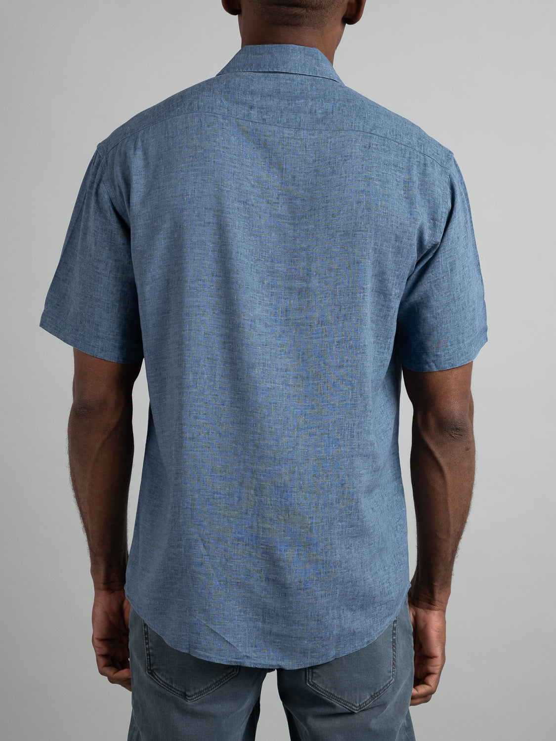Rear view of a person wearing the Maharishi Rayon Linen Short Sleeve Shirt by ID Menswear in blue with gray pants. The rayon linen blend fabric ensures comfort and style, set against a neutral background.