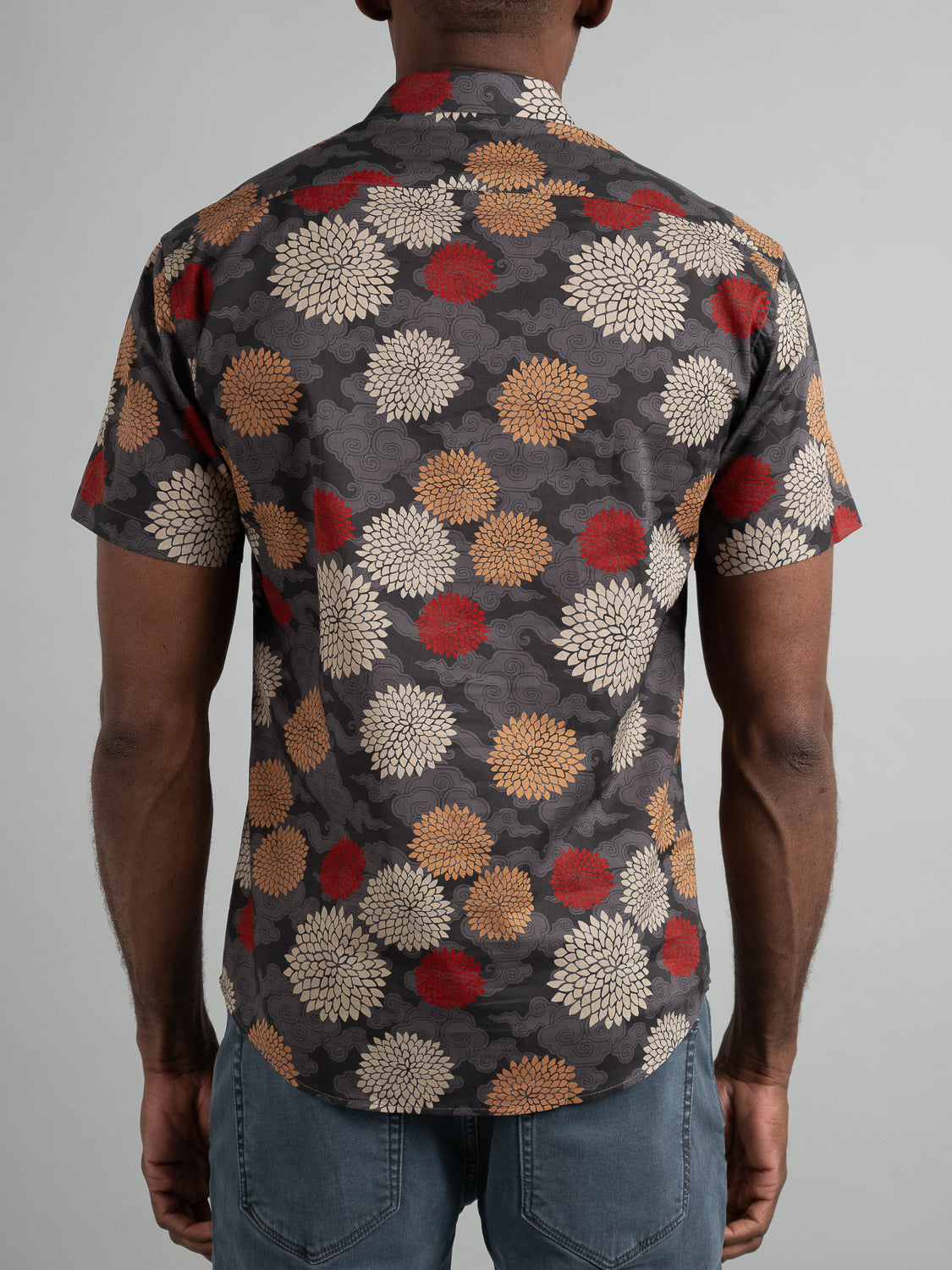 Someone is wearing a Tibet Short Sleeve Cotton Printed Shirt by ID Menswear, featuring large, bold red, beige, and gray flowers on a plain gray background with their back to the camera.