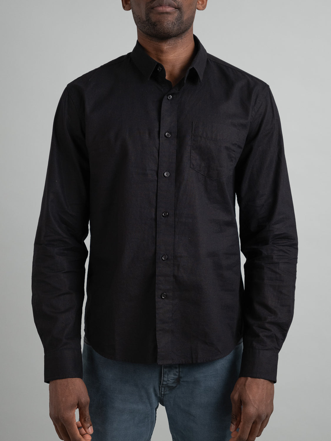 A person in a lightweight ID Menswear Goa Linen Cotton Long Sleeve Shirt, black with a collar and chest pocket, pairs it with blue jeans against a plain light gray background.