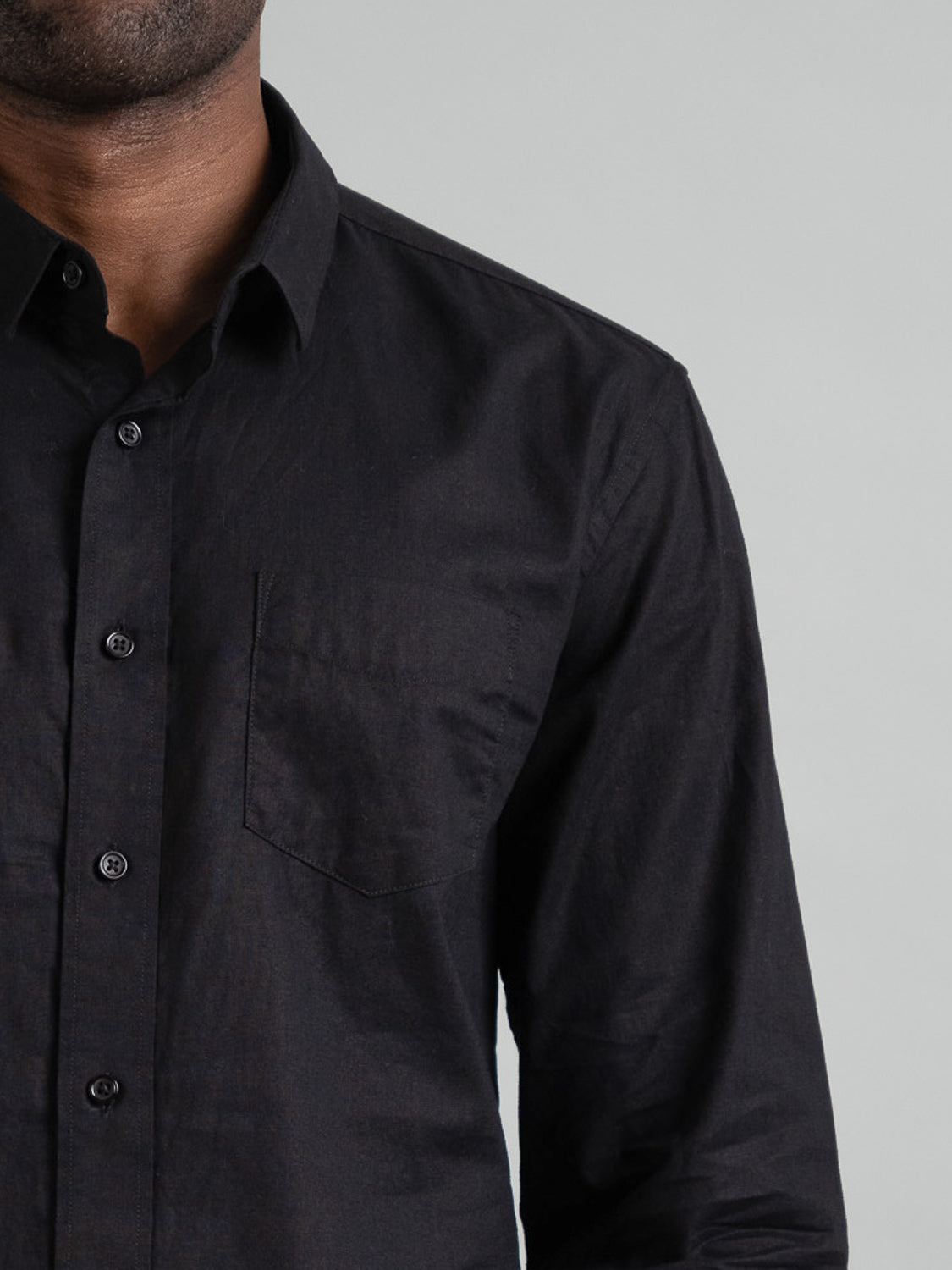 A person models their upper body in a black Goa Linen Cotton Long Sleeve Shirt by ID Menswear, featuring a button-up design and front pocket, against a plain gray background.