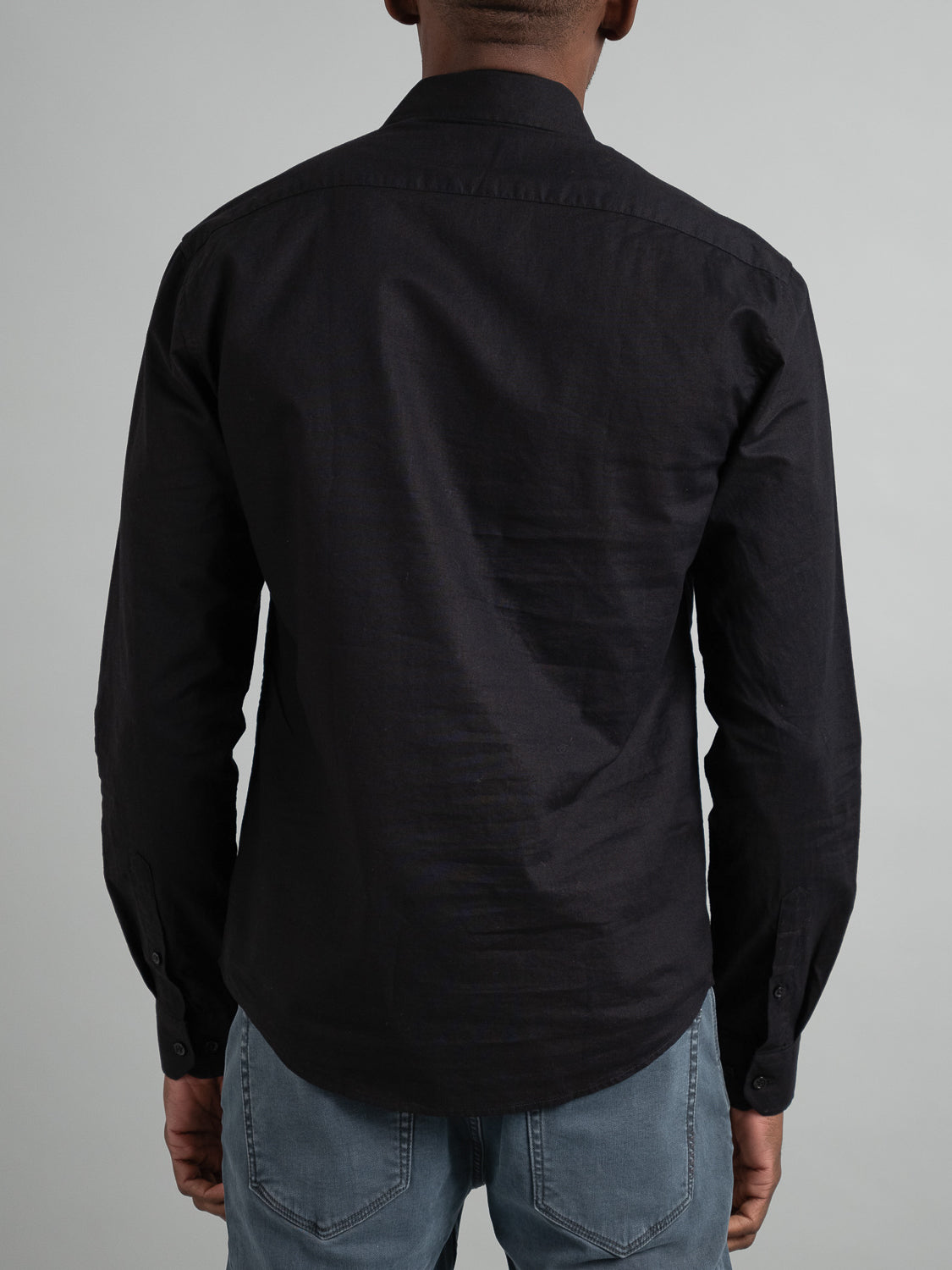 A person is seen from behind wearing the Goa Linen Cotton Long Sleeve Shirt in black by ID Menswear. Its lightweight, breathable fabric flows gracefully against a plain gray backdrop, paired with light blue jeans.