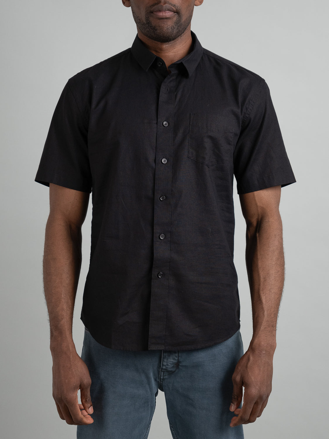 A person in a black ID Menswear Ravi Linen Cotton Short Sleeve Shirt with a chest pocket and blue jeans stands against a light gray background.