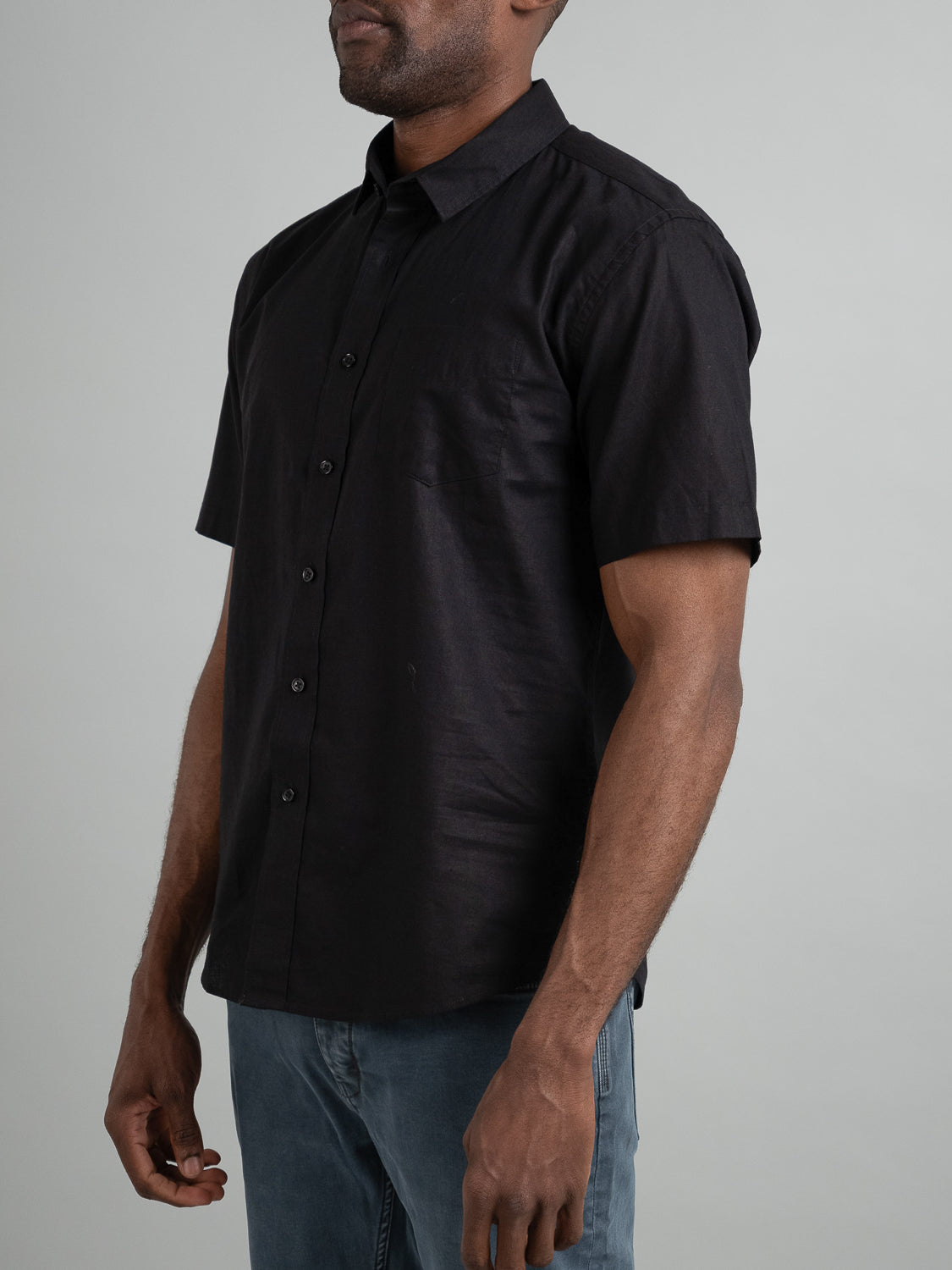 A man in a Ravi Linen Cotton Short Sleeve Shirt by ID Menswear, featuring a breathable black linen-cotton blend, paired with blue jeans, stands against a plain gray background with his head not visible in the image.