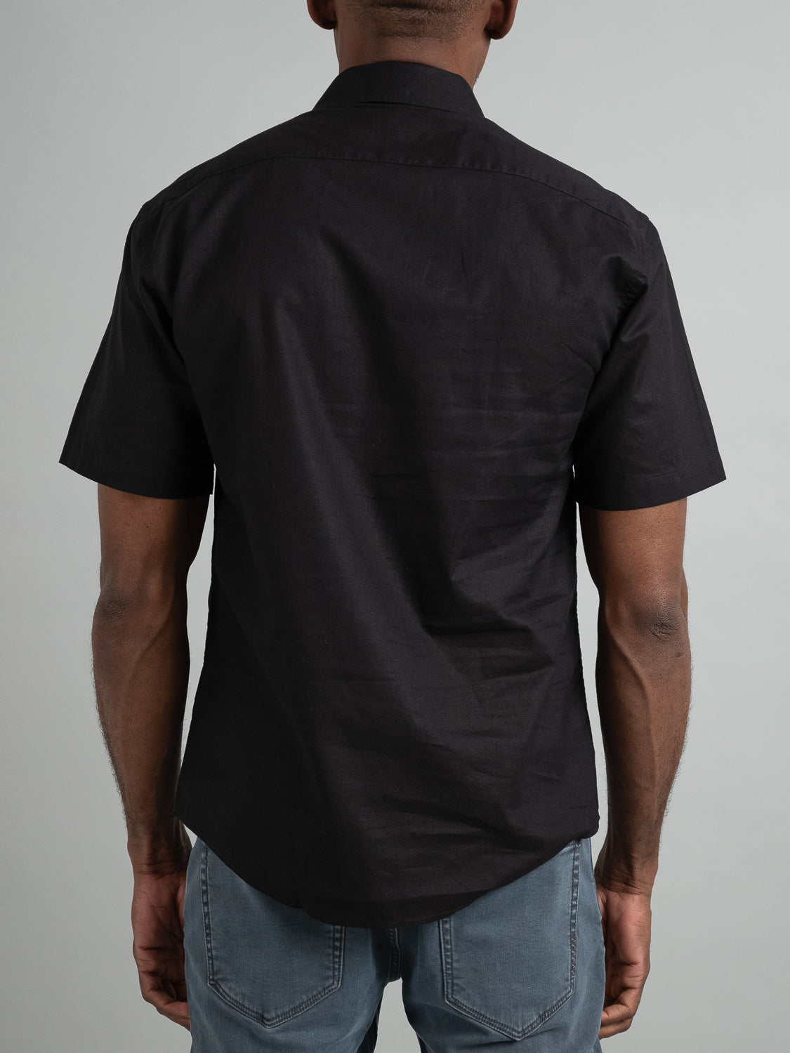 A man, back turned, wears the Ravi Linen Cotton Short Sleeve Shirt from ID Menswear, a black button-up made of breathable linen-cotton blend, paired with light blue jeans against a gray backdrop.