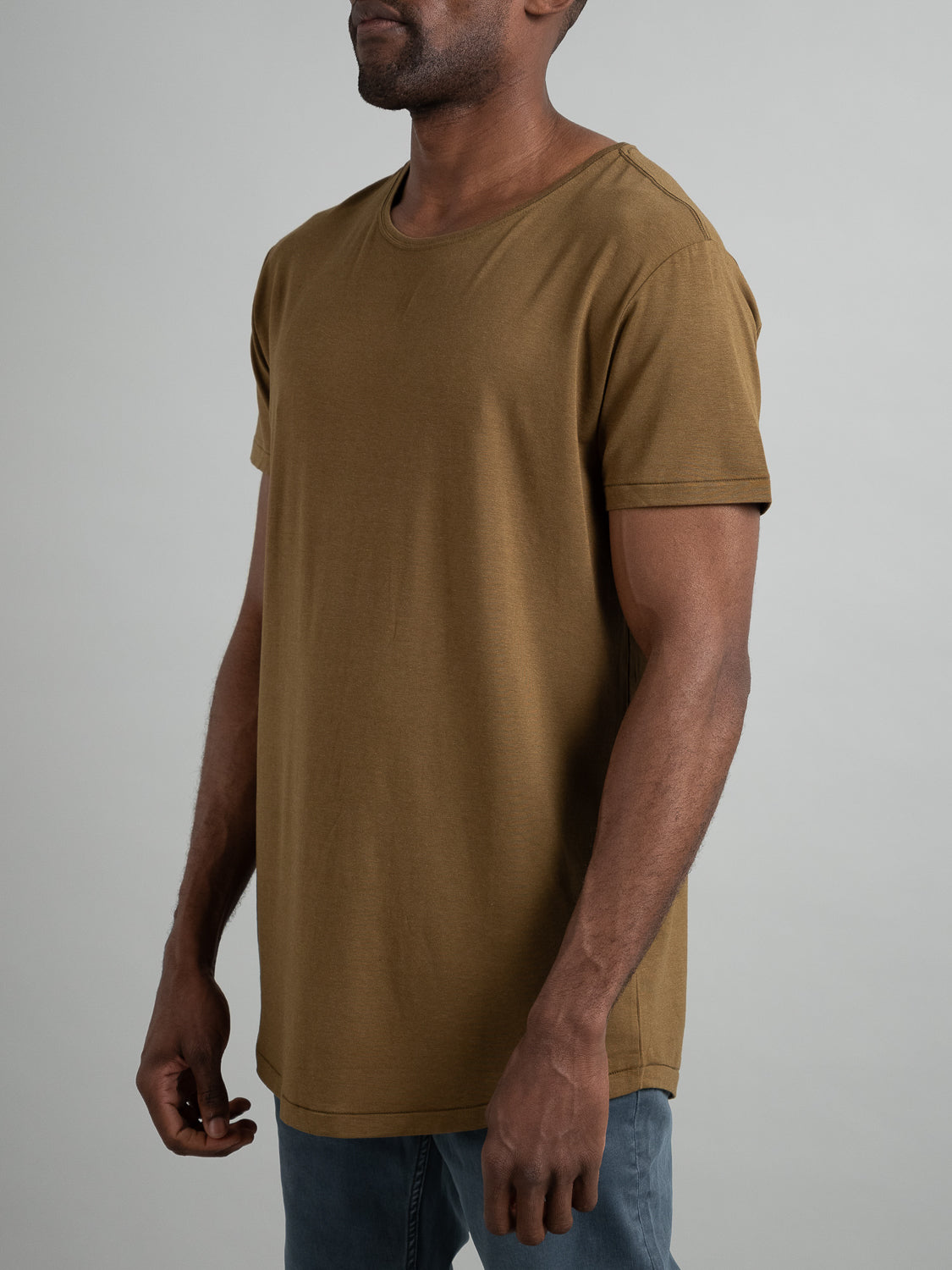 Against a neutral gray background, a model wearing an olive Slay Wide Neck Bamboo Blend Tee from ID Menswear stands side-facing with relaxed arms, paired with light blue jeans crafted from soft organic cotton.